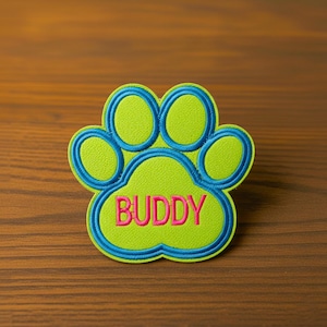 Op de afbeelding: Geborduurde patch in de vorm van een hondenpootafdruk. De poot is limoengroen met een blauwe omtrek en het woord "BUDDY" in roze. De patch ligt op een houten oppervlak.