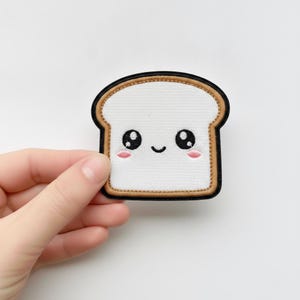 Puede incluir: Un parche bordado con forma de rebanada de pan blanco con un borde de corteza tostada y un contorno negro. El centro presenta una cara de dibujos animados con dos grandes ojos negros, una pequeña sonrisa negra y dos círculos rosados.
