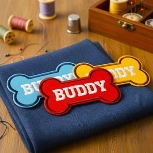 Può includere: Tre toppe ricamate a forma di osso, blu, rosse e gialle, ciascuna con il nome "BUDDY" a lettere bianche. Le toppe sono su un tessuto blu, con forniture di cucito sullo sfondo.