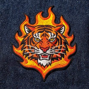 Pode incluir: Patch bordado com a cabeça de um tigre em chamas. O tigre tem listras laranja e pretas, olhos amarelos e dentes brancos. As chamas são em tons de laranja e amarelo. O patch está em um fundo de ganga azul escuro.