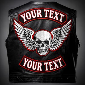 May include: A black leather vest featuring a large embroidered patch on the back. The patch displays a white skull with expansive wings, flanked by two curved banners with red borders that contain the white text YOUR TEXT.