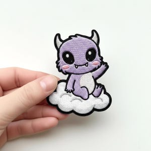 Peut inclure: Écusson brodé représentant un monstre violet de dessin animé avec des cornes noires, de grands yeux et un ventre blanc. Le monstre est assis sur un nuage blanc avec une bordure noire, faisant un signe de la main. Pour vêtements ou accessoires.