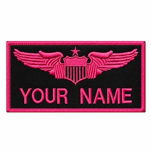 Op de afbeelding: Een rechthoekige zwarte geborduurde patch met een felroze rand. Het ontwerp toont een centraal schild met verticale strepen, bekroond door een ster en geflankeerd door twee vleugels. De tekst YOUR NAME is in het roze onder het embleem geborduurd.