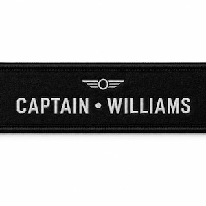 Könnte beinhalten: Ein rechteckiger, schwarz bestickter Aufnäher mit weißem Rand. Der Text CAPTAIN • WILLIAMS ist in weißen Großbuchstaben zentriert. Über dem Text befindet sich ein kleines, weiß gesticktes Emblem mit einem Mittelkreis und zwei Flügeln.