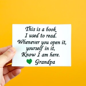 Puede incluir: Una tarjeta rectangular blanca con texto negro que dice "This is a book I used to read. Whenever you open it, yourself in it, Know I am here. Grandpa." Un corazón verde está debajo del texto. La tarjeta se sostiene contra un fondo amarillo.