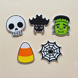 May include: A collection of five Halloween-themed embroidered patches. Includes a white skull with pink cheeks, a black bat, a green Frankenstein's monster, a candy corn, and a black and white spiderweb.