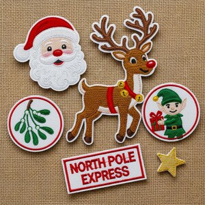 Pode incluir: Patches de Natal bordados com o Pai Natal, uma rena com sinos, um duende, azevinho, uma estrela e um letreiro com a frase "North Pole Express". Os patches são nas cores vermelho, branco, castanho e verde.