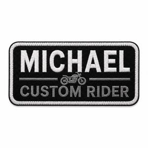 Op de afbeelding: Een rechthoekige geborduurde patch met een zwarte achtergrond en een witte rand. De tekst MICHAEL staat in het wit bovenaan, gevolgd door een klein grijs motorpictogram en de tekst CUSTOM RIDER in het grijs onderaan de patch.