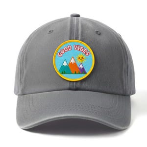 May include: Grey baseball cap with a curved peak. The cap has a circular patch with the words "GOOD VIBES" around a cartoon image of mountains and a smiling sun.