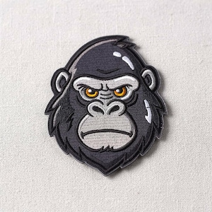 Puede incluir: Parche bordado con la cabeza de un gorila. El gorila tiene una expresión seria con ojos naranjas, hocico gris y pelaje negro. El parche está delineado con hilo negro y blanco, dándole un aspecto de dibujos animados. Para ropa o accesorios.