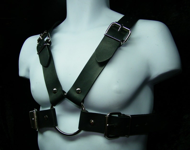 Op de afbeelding: Een zwart leren harnas met zilveren gespen en een grote metalen ring. Het harnas wordt gedragen door een mannequin.