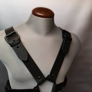 Op de afbeelding: Een zwart leren harnas met een metalen ring en gespen. Het harnas wordt getoond op een grijze mannequin.