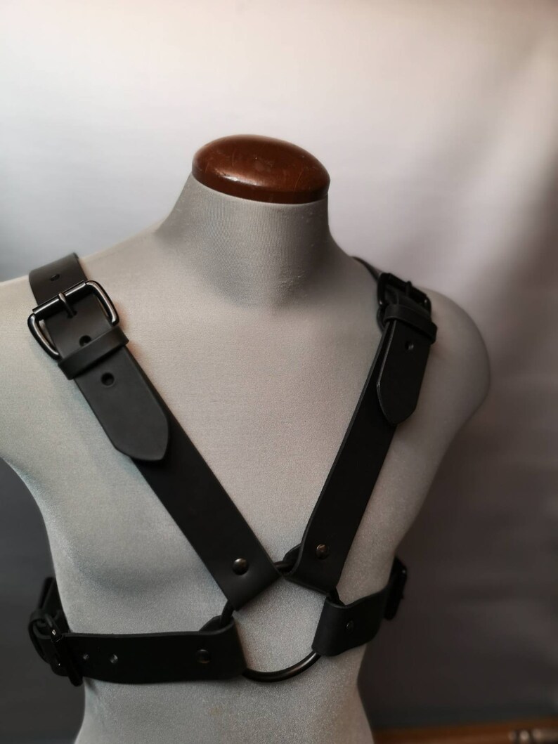 Op de afbeelding: Een zwart leren harnas met een metalen ring en gespen. Het harnas wordt getoond op een grijze mannequin.