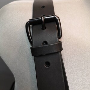 Op de afbeelding: Zwarte leren riem met een zwarte metalen gesp. De riem wordt getoond op een wit mannequin.