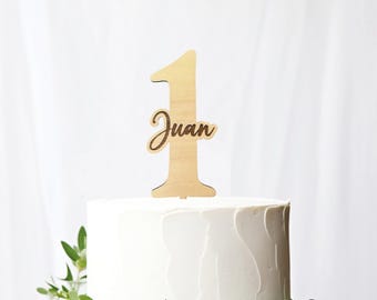 May include: A wooden cake topper in the shape of the number 1 with the name "Juan" in script. The topper is placed on top of a white frosted cake decorated with greenery. The background is a plain white.