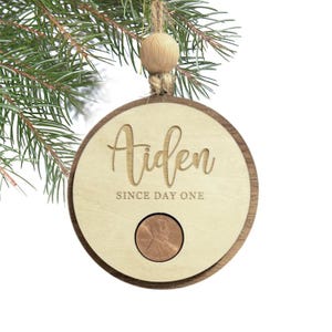 May include: Wooden Christmas ornament with the name "Aiden" engraved, along with "SINCE DAY ONE". A penny is set into the ornament. The ornament is hanging from a twine string and wooden bead.