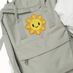 May include: A light gray backpack with a yellow smiling sun patch. The backpack has a front pocket with a zipper and adjustable straps. The sun patch is yellow with an orange outline and a smiling face.