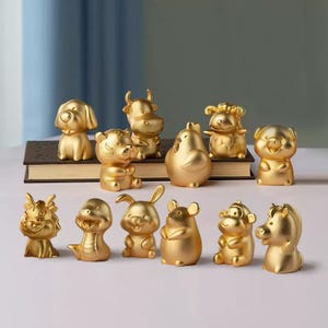 May include: A collection of twelve small, gold-colored animal figurines representing the Chinese zodiac. The set includes a dog, ox, tiger, rooster, sheep, pig, dragon, snake, rabbit, rat, monkey, and horse, all arranged in two rows.