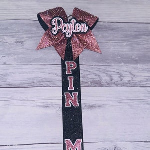 May include: A black and pink cheer bow with the name "Peyton" on it. The bow is attached to a black ribbon with the words "PIN ME" written in pink letters.