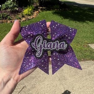 May include: A purple glitter bow with rhinestones and the name "Gianna" written in silver glitter on a black background.
