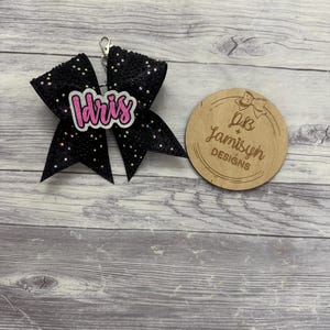May include: A black glitter bow with the name "Idris" in pink and white lettering. The bow has a silver clasp and is next to a wooden circle with the text "PB Jamisyn Designs". The background is a light grey wood grain.