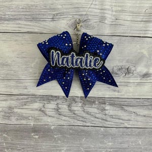 Puede incluir: Un lazo de animadora azul y plateado con purpurina, con un llavero plateado y el nombre "Natalie" escrito en él.