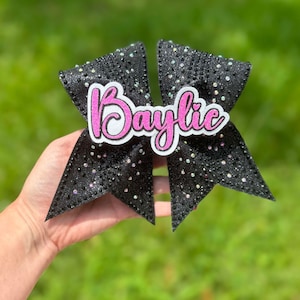 May include: Black cheer bow with iridescent rhinestones and a pink glitter name "Baylie" on the front.