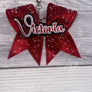 May include: Red glitter cheer bow with a black and white name tag that says "Victoria" and a silver keychain attached.