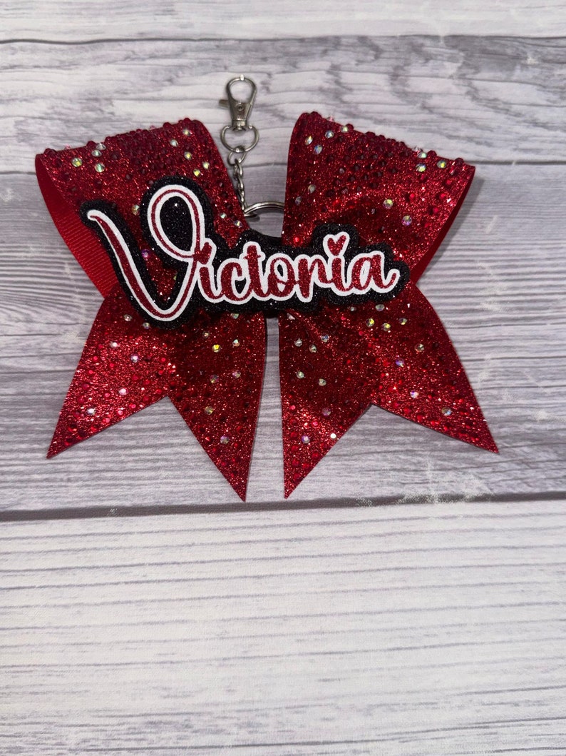 May include: Red glitter cheer bow with a black and white name tag that says "Victoria" and a silver keychain attached.