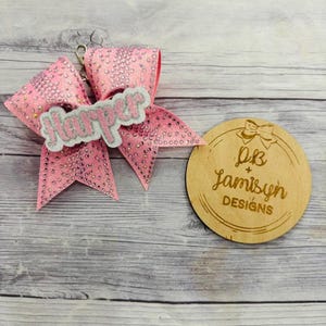 Pode incluir: Um laço de animadora rosa e branco com o nome "Harper" em letras prateadas com glitter. O laço é decorado com pedrarias. Um círculo de madeira com o texto "JB + Jamisyn DESIGNS" está no fundo.