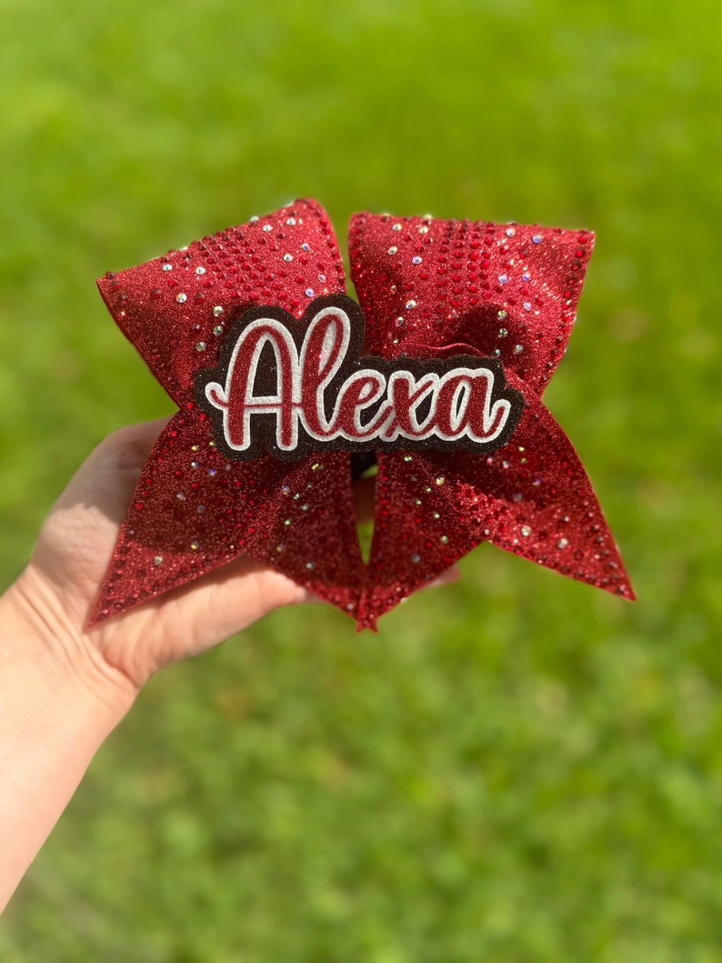 May include: A red glitter cheerleading bow with the name "Alexa" in white script with a black and white border. The bow is held up against a green background.