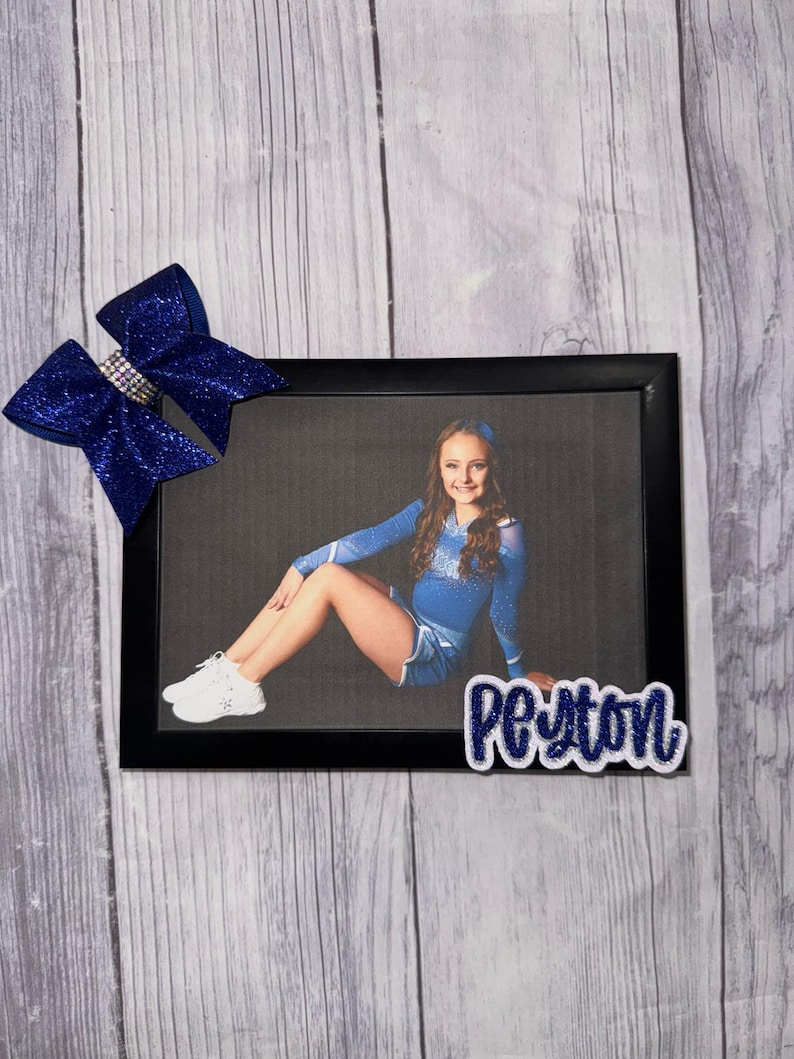 May include: A black picture frame with a photo of a young girl in a blue cheerleading uniform. The girl is sitting on the floor with her legs crossed and her arms around her knees. The frame has a blue and white glitter bow on the top left corner and a white glitter name tag that says "Peyton" on the bottom right corner.