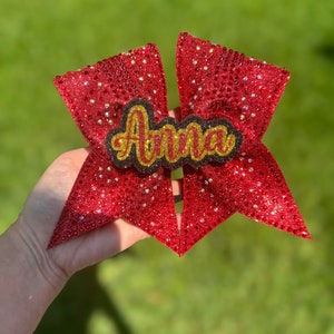May include: A red glitter cheerleading bow with the name "Anna" in gold lettering. The bow is held against a green background. The bow is covered in small, sparkling embellishments.