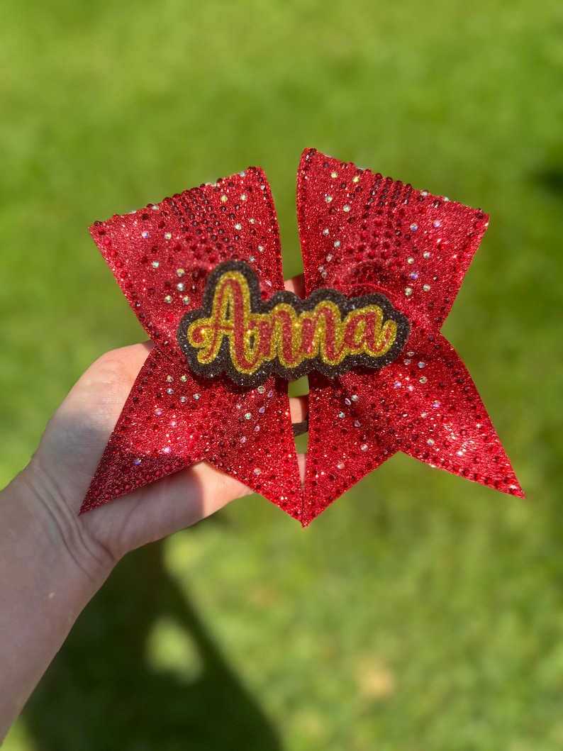 May include: A red glitter cheerleading bow with the name "Anna" in gold lettering. The bow is held against a green background. The bow is covered in small, sparkling embellishments.