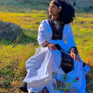 Puede incluir: Una mujer vestida con un vestido tradicional etíope blanco con bordados coloridos y una diadema marrón. Está sentada en un campo de hierba con una gran roca detrás de ella.