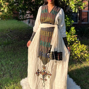 Puede incluir: Vestido color crema de manga larga con intrincados bordados multicolores. El vestido presenta un cuello en V, un cinturón envolvente y patrones detallados en el corpiño y las mangas. El vestido se usa sobre una alfombra de piel sintética blanca.