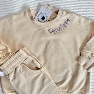 May include: A beige two-piece outfit for children, including a sweatshirt and sweatpants. The sweatshirt has the name "Penelope" embroidered in blue thread. The Black Sheep Boutique tag is attached. The outfit is made of soft material.