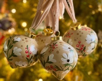 May include: Four beige Christmas ornaments with gold caps and cream-colored floral designs, including white flowers and red berries. The ornaments are suspended by beige ribbons against a blurred background of a Christmas tree.