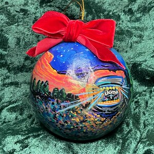 May include: A hand-painted Christmas ornament with a vibrant landscape scene, featuring a sunset, trees, and the word "ZACHTOP". The ornament is topped with a red velvet bow and gold string for hanging.