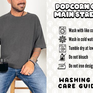 Puede incluir: Una camiseta gris oscuro con el texto "Popcorn on Main Street" impreso en ella. La imagen tambi&eacute;n incluye una gu&iacute;a de lavado y cuidado con instrucciones para lavar con colores similares, lavar con agua fr&iacute;a, secar en secadora a baja temperatura, no usar lej&iacute;a y no planchar el dise&ntilde;o.