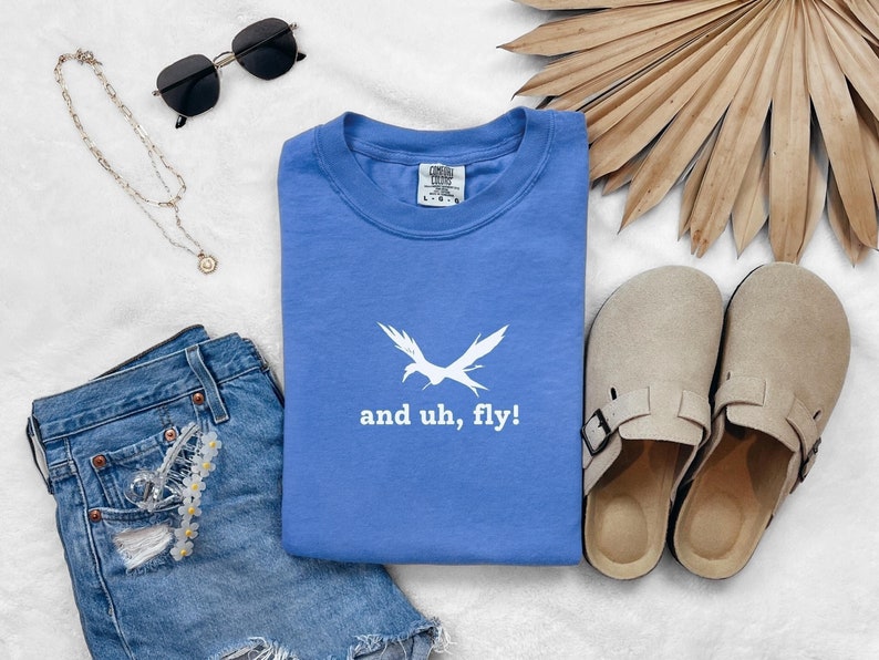 Puede incluir: Una camiseta azul con un gr&aacute;fico de p&aacute;jaro blanco y el texto "and uh, fly!"