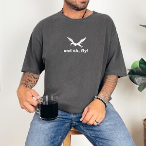 Puede incluir: Una camiseta gris oscuro con un gr&aacute;fico de p&aacute;jaro blanco y el texto "and uh, fly!"