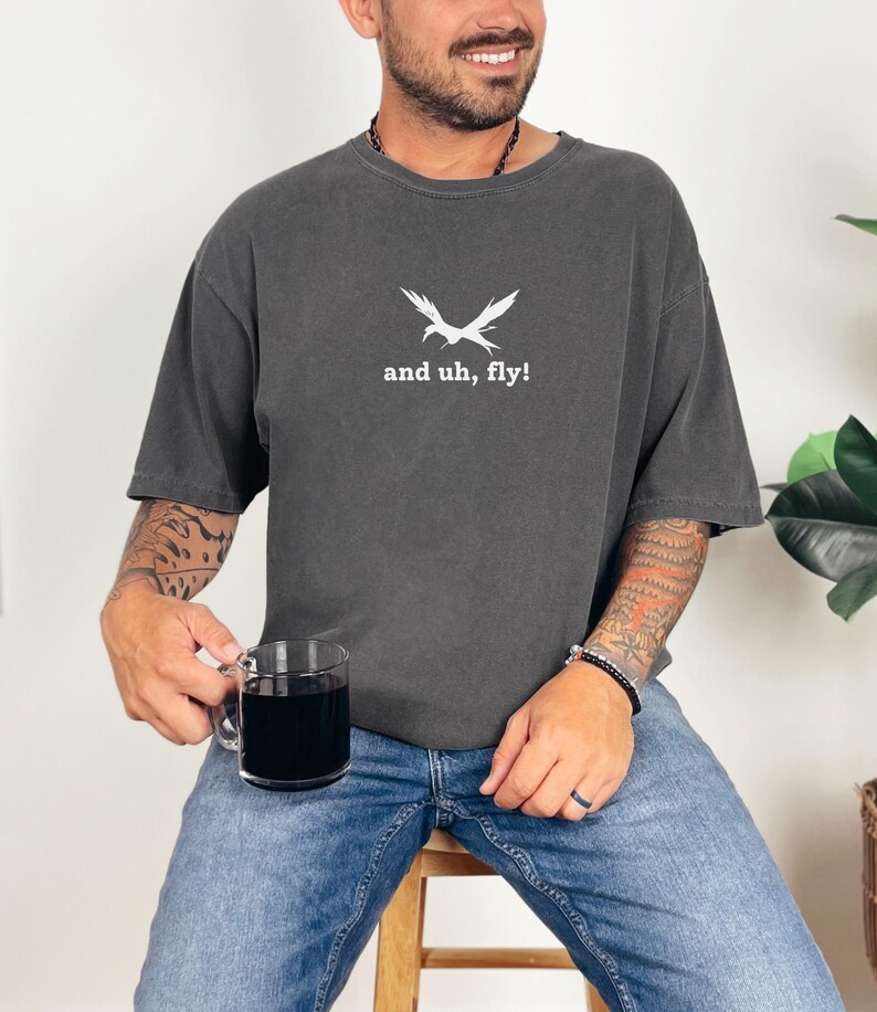 Puede incluir: Una camiseta gris oscuro con un gr&aacute;fico de p&aacute;jaro blanco y el texto "and uh, fly!"
