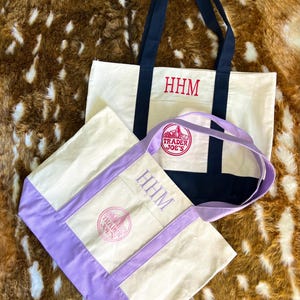 May include: Two canvas tote bags featuring embroidered monograms and logos. One bag has navy blue handles and base with red HHM text and a Trader Joe's logo. The other bag has lavender handles and base with lavender HHM text and a Trader Joe's logo.