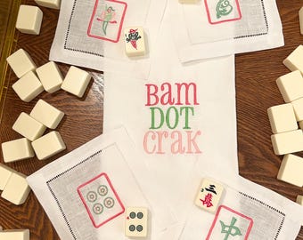 May include: White linen napkins with embroidered red and green designs, and the text "bam dot crak". The napkins are surrounded by ivory Mahjong tiles, creating a game-themed arrangement.