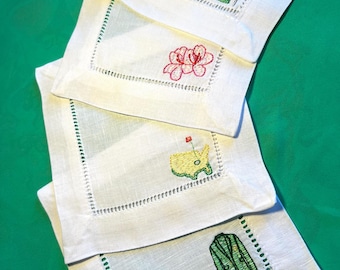 May include: White linen coasters with embroidered designs. Each coaster features a different design: a building with a flag, a pink flower, a golf course map, and a green blazer. The coasters have a green stitched border.