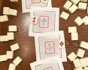 May include: A collection of ivory-colored Mahjong tiles and four white linen coasters with embroidered Mahjong tile designs in red and green. The coasters have a black stitched border. The tiles are scattered on a wooden surface.