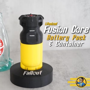 May include: Lifesized Fallout Fusion Core Battery Pack & Container. The yellow and black battery pack is on a black stand with the word "Fallout" printed on it. The battery pack has a black top with a red button.