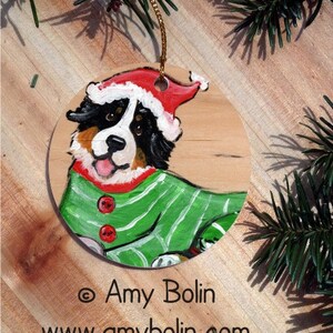 May include: A wooden Christmas ornament featuring a Bernese Mountain Dog wearing a Santa hat and green striped pajamas. The ornament has a gold string for hanging and is painted with red, white, black, and brown colors.