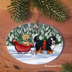 May include: A Christmas ornament featuring a Bernese Mountain Dog pulling a red sled with a Golden Retriever puppy inside. The dogs are in a snowy forest setting with green trees and white snow.